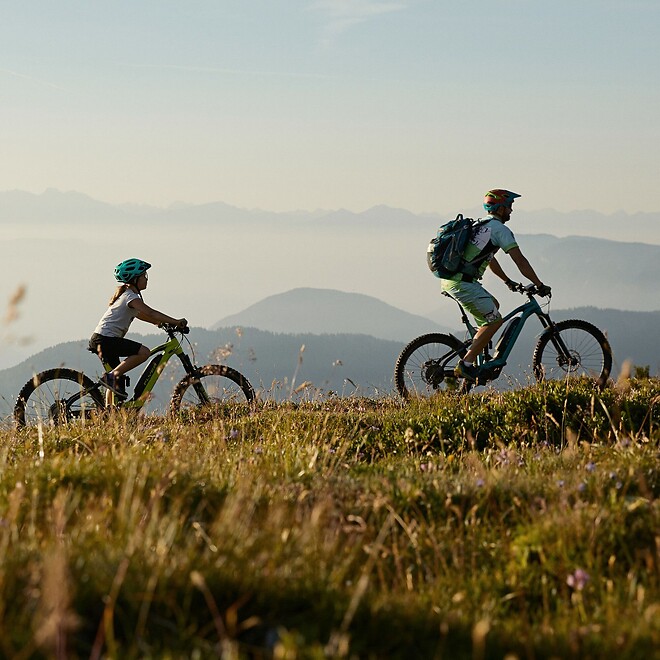 Familie fährt mit E-Bikes über befestigtem Bergweg vor Bergpanorama und Abendsonne