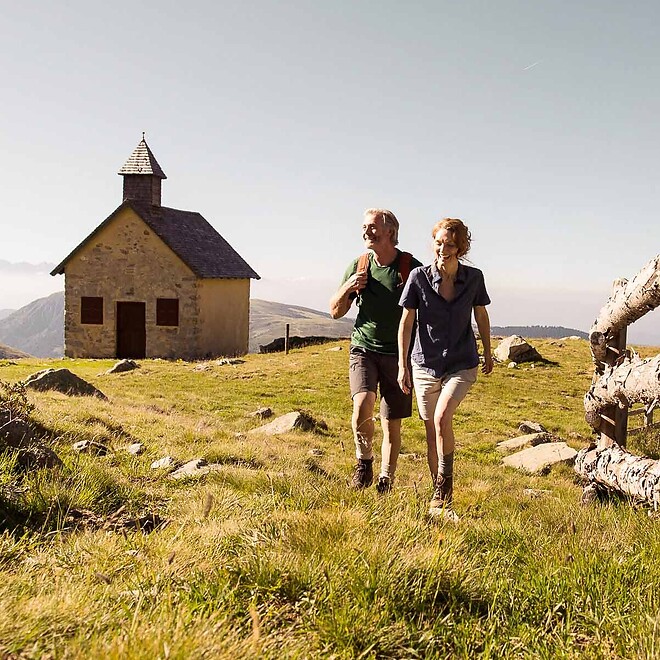 Man and woman hiking across meadow past small church in the mountains
