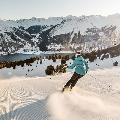 A skier carves down a freshly groomed slope; in the background, a wide panoramic view of Lake Reschen and the snow-capped peaks of the Alps unfolds under bright sunshine.