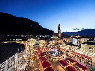 An atmospheric aerial evening shot of the Bolzano Christmas Market, featuring illuminated stalls on Walther Square set against the silhouette of the Bolzano Cathedral and the surrounding mountains.