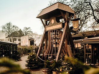 A large, rustic wooden bell tower at the Forst Christmas Forest in Algund, surrounded by festive Christmas decorations and trees under a clear sky.