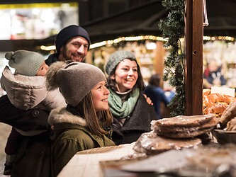 A family in warm winter clothing stand at a cozy, illuminated Christmas market stall and look at the goods on display.