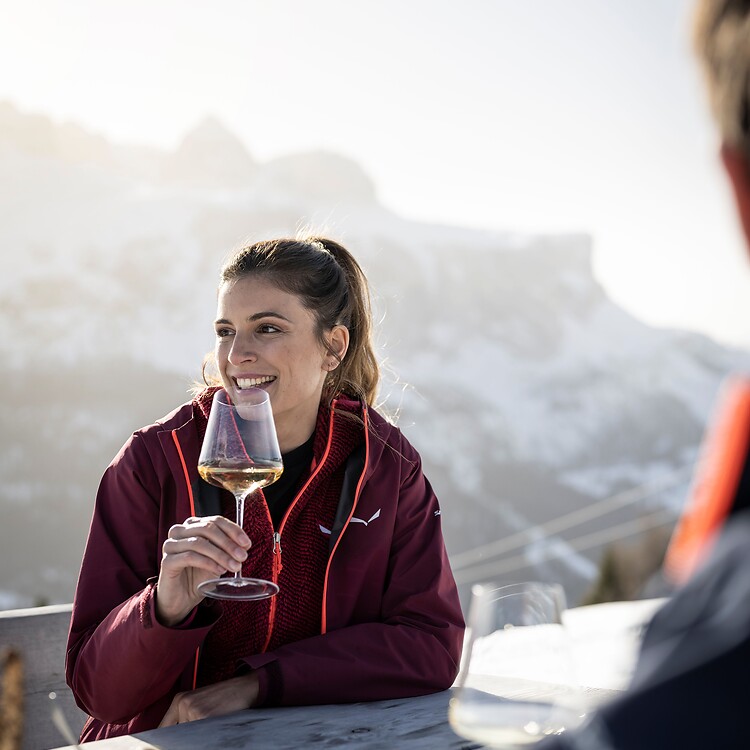 A smiling woman holding a glass of white wine on a sunny terrace overlooking the snow-covered Dolomites.